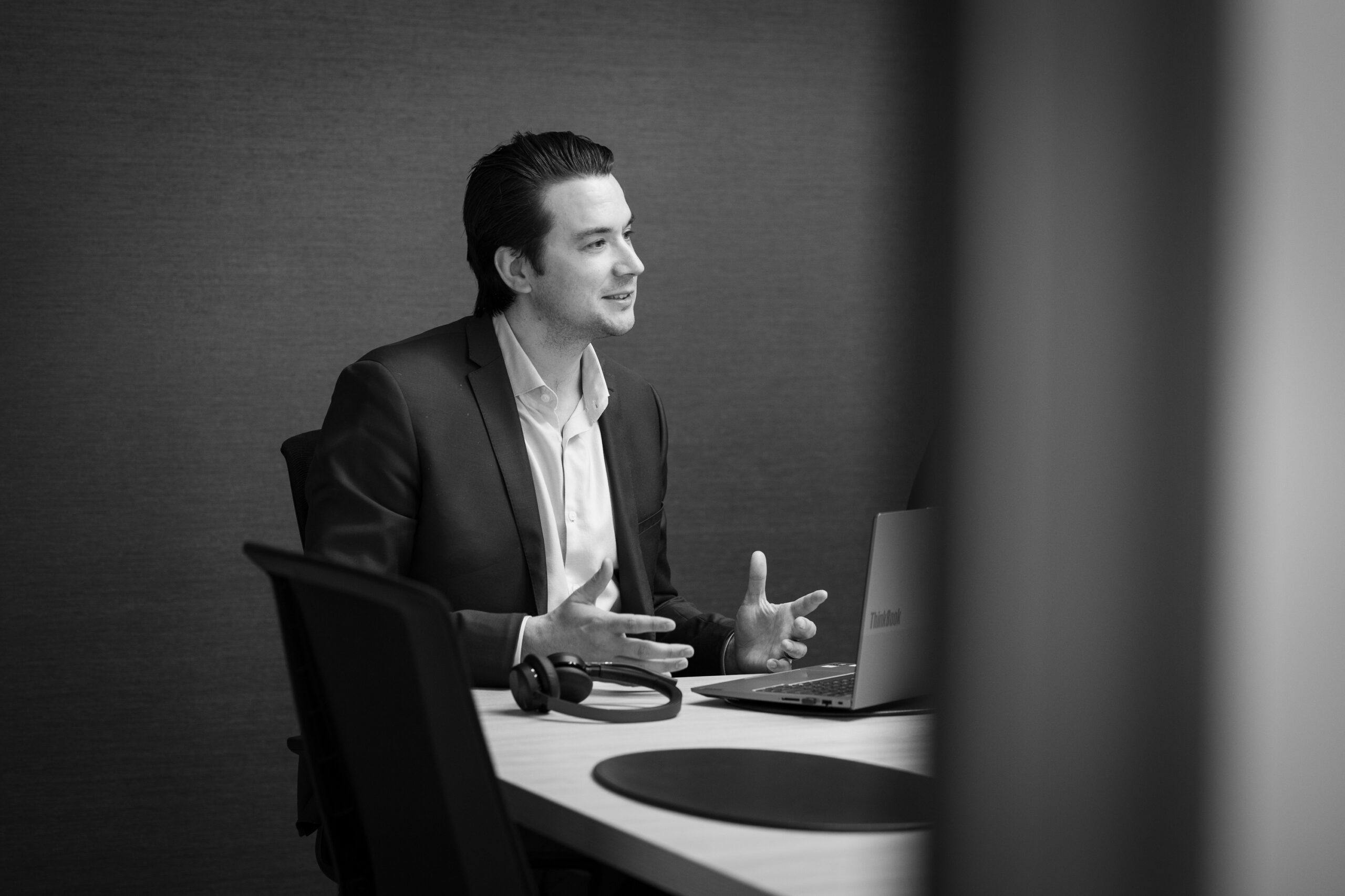 A young man in a suit sits at a table with a lapotp and headphones.