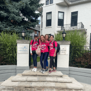 Four women in matching pink shirts, leggings and running shoes stand together outside a homey white office building.