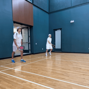 Two men in white shirts and shorts playing pickleball indoors.