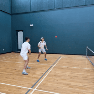 Two men in white shirts and shorts playing pickleball indoors.