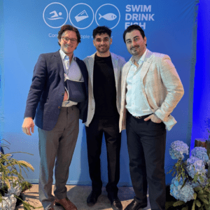 Three men in business casual attire standing together and smiling in front of a blue Swim, Drink, Fish backdrop
