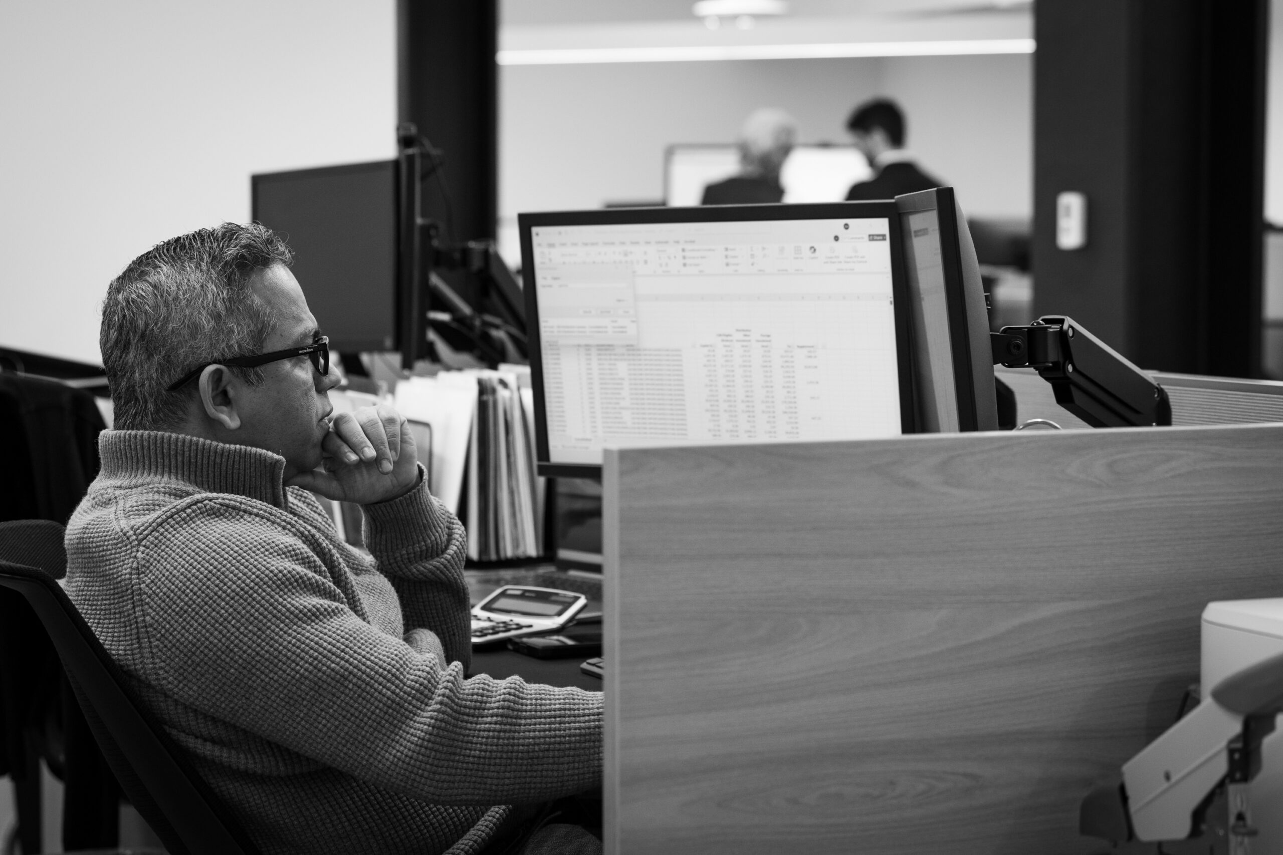 A man in a sweater and glasses sitting at a desk looking at a large monitor.
