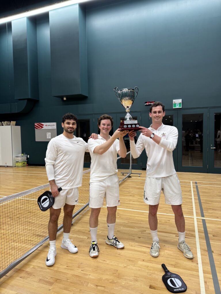Three young men in white pickleball outfits old a silver trophy while standing on a pickle ball court.