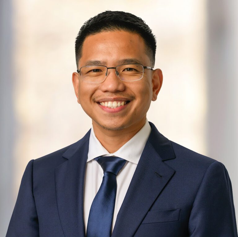 A man wearing glasses and a navy blue suit, white dress shirt, and dark blue tie, posing for a headshot.